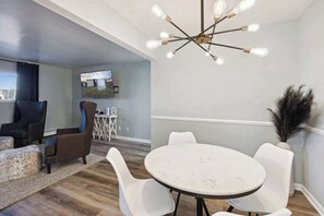 A modern living room with a round table, white chairs, a brown armchair, a television, and a unique ceiling light fixture. - Lansing Layover#3 | 1 BR - 1 BA (Lansing)