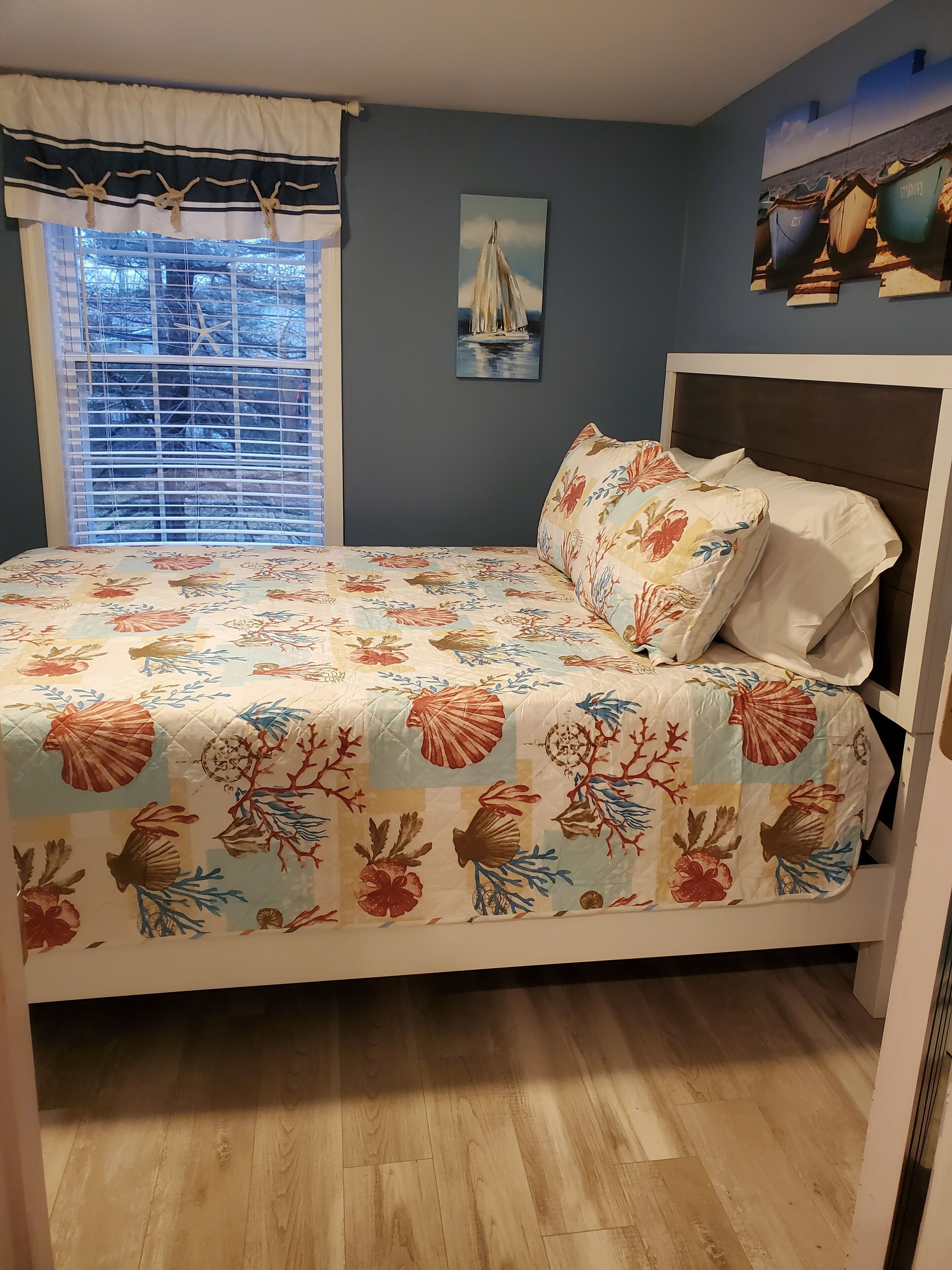 A bedroom with a bed, a window with blinds, a wall with a painting, and a wooden floor.