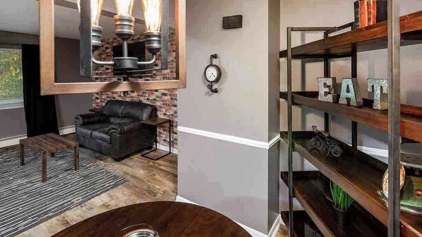 Una sala de estar moderna con un sofá de cuero, una mesa de centro redonda, una pared decorada con ladrillos, un reloj de pared y una estantería de metal con la palabra 'EAT'.