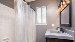 Un baño con cortina de ducha, una ventana con persianas, un lavabo con espejo y una lámpara.
