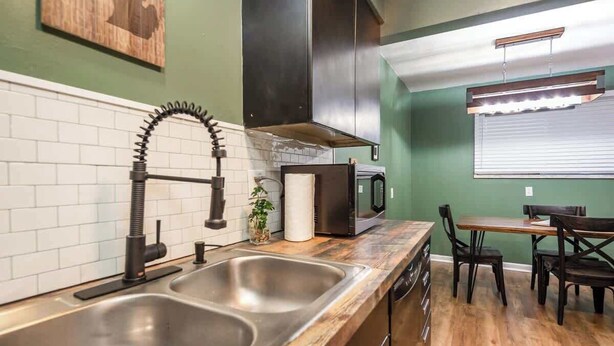 A modern kitchen with a stainless steel sink, a black faucet, a microwave, and a wooden dining table with chairs. - Fairlane #8 | The Den (Lansing)