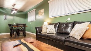 A living room with a black leather sofa, wooden coffee table, and a dining area with a wooden table and chairs. - Fairlane #8 | The Den (Lansing)