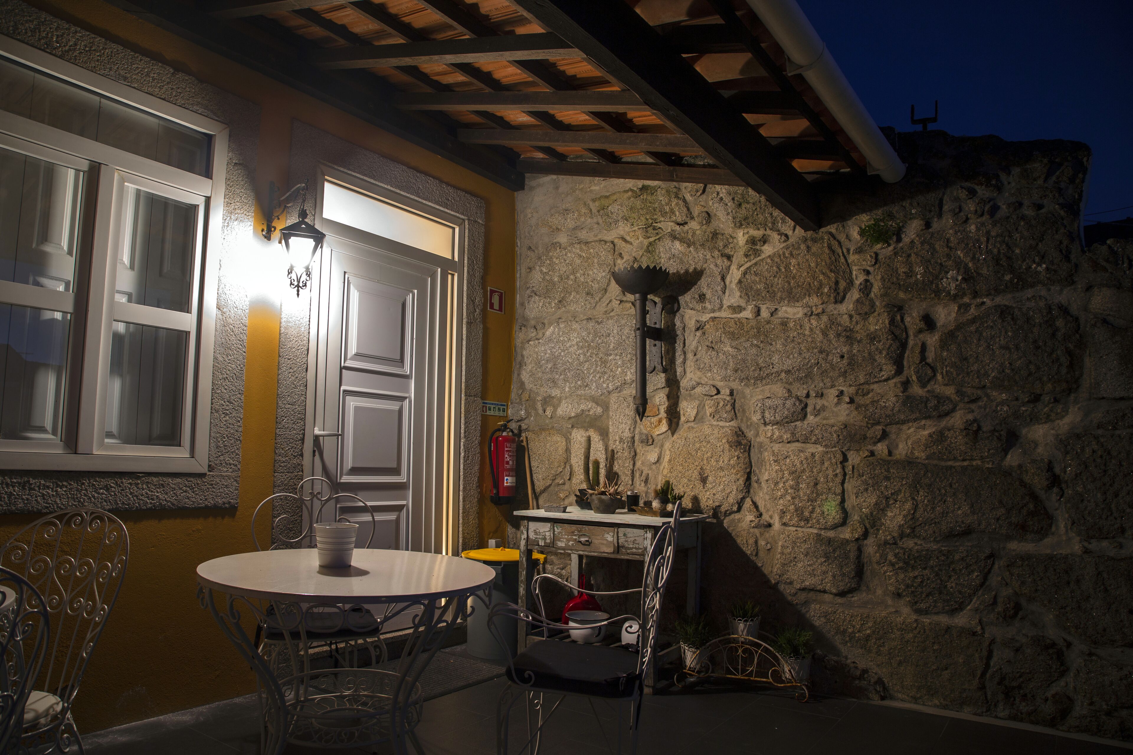 Un muro de piedra con un farol de metal, una puerta blanca y una mesa redonda con sillas.