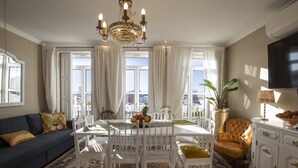 A dining area with a white table, chairs, and a chandelier. There is a sofa with a lion pillow and a plant in the corner.