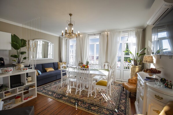 A living room with a dining table, white chairs, a chandelier, and a large window with curtains.
