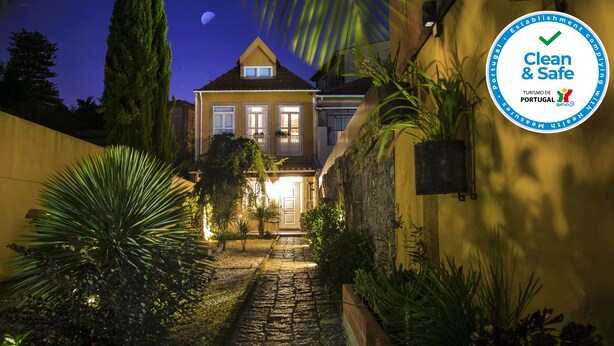 A well-lit pathway leading to a house with a balcony, surrounded by various plants and a stone pathway.