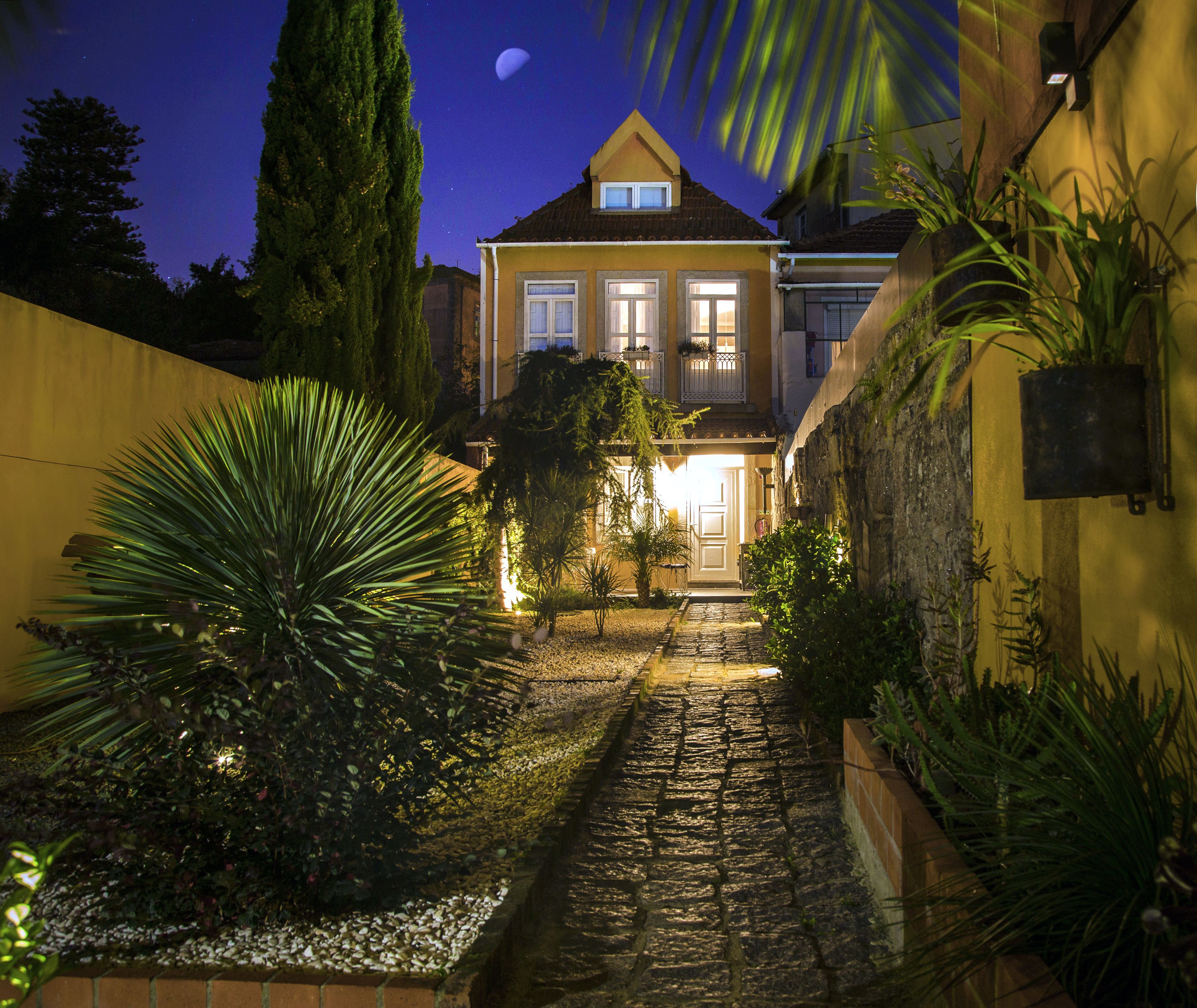 Un chemin bien éclairé mène à une maison dotée d’un balcon, qui est entourée de différentes plantes et bordée d’un sentier en pierre.