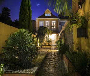 A well-lit pathway leading to a house with a balcony, surrounded by various plants and a stone pathway.
