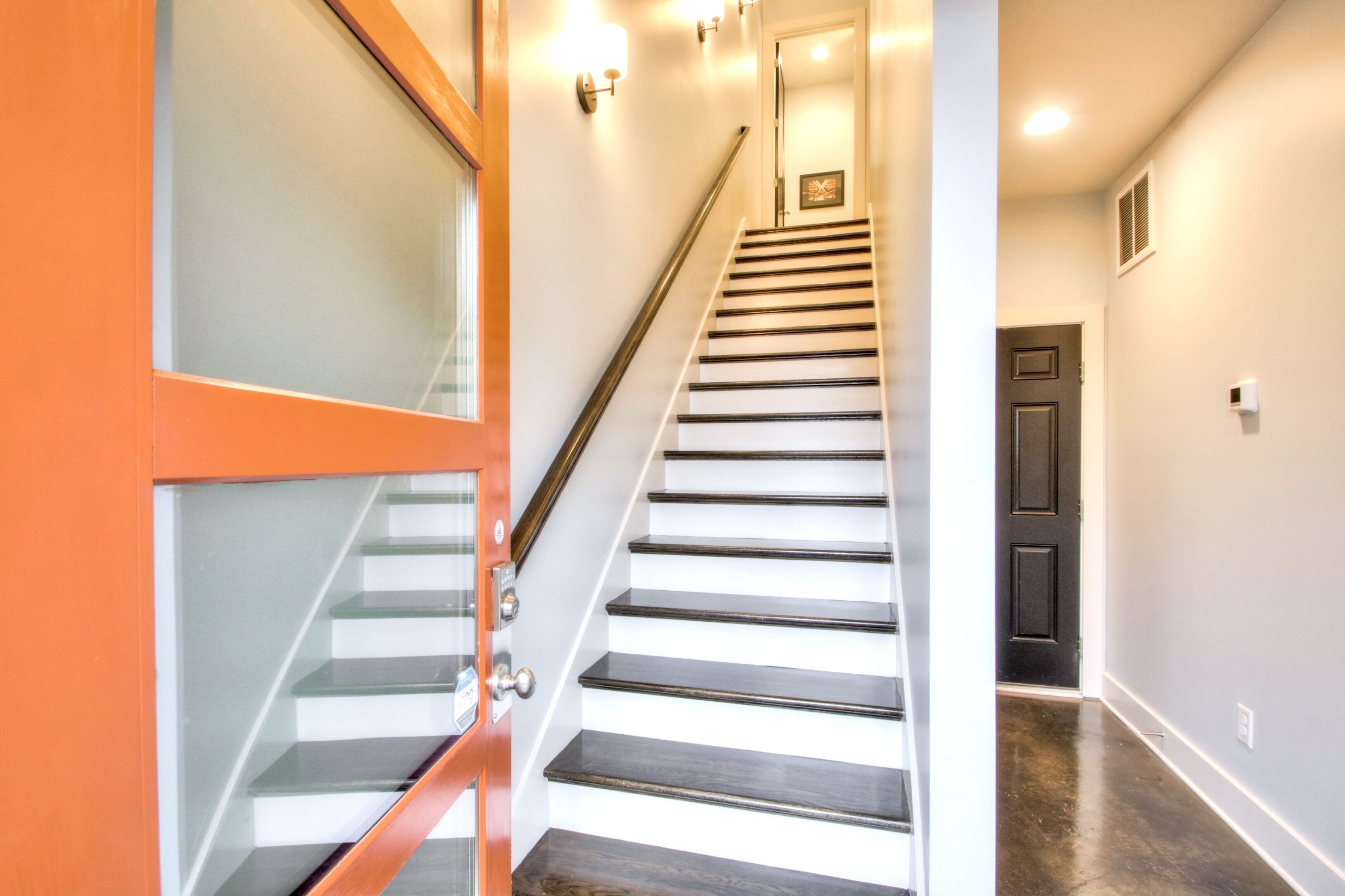 A staircase with a wooden handrail and glass panels on the left, leading to a dark door at the top.