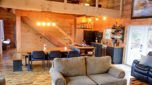 Interior acogedor de una cabaña con escalera de madera, comedor, cocina y sala de estar con sofá y chimenea.