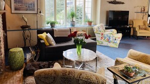 A cozy living room with a sofa, armchairs, a coffee table, and a TV. There are potted plants and a large window providing natural light. - Lg Victorian, New Resort-likePrivate Pool, Beds for 17, 10min to SH beach (Cape May Court House)