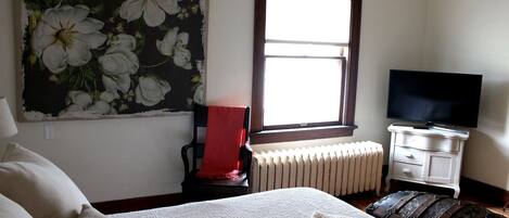 A bedroom with a bed, a chair, a television, a radiator, and a window.