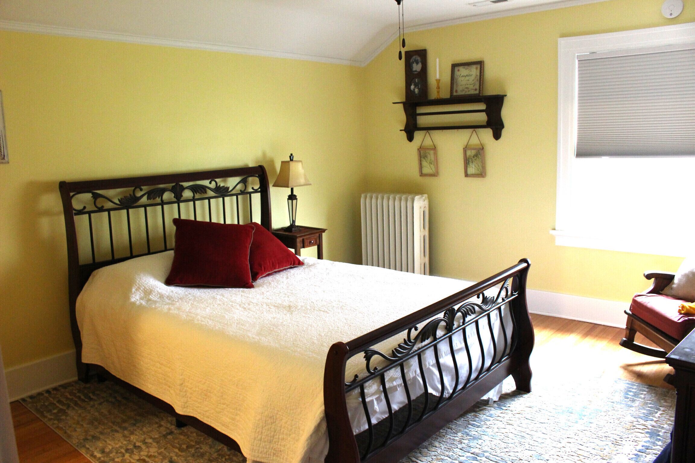 Une chambre à coucher comprenant un lit, une table de chevet, une lampe, un radiateur et une fenêtre avec des stores.