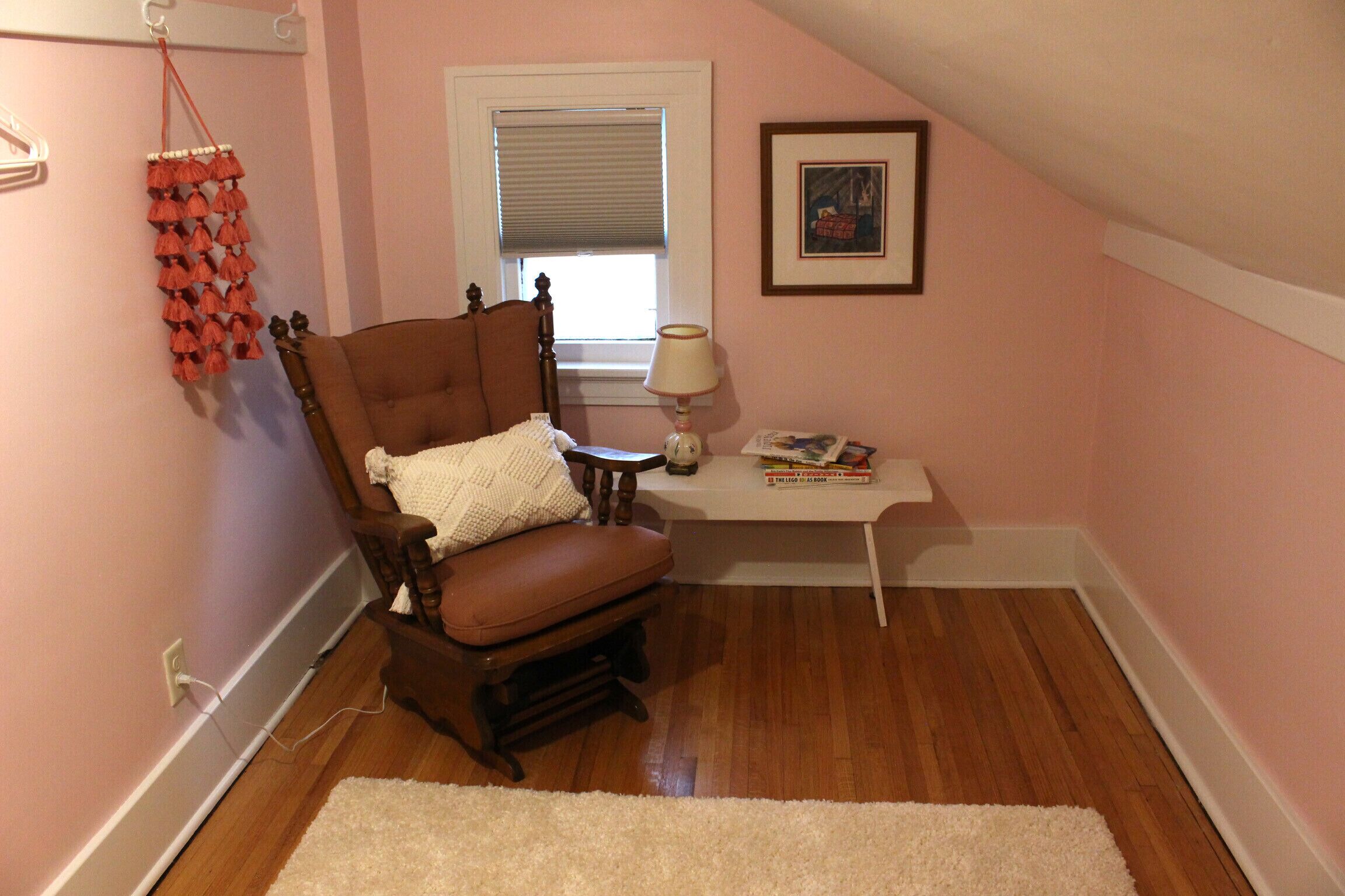 A cozy room with a wooden chair, a white rug, a small table with a lamp, and a window with blinds.