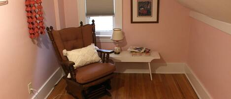 A cozy room with a wooden chair, a white rug, a small table with a lamp, and a window with blinds.