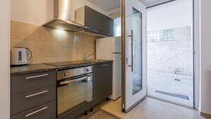 Cocina compacta con estufa, horno y hervidor, puerta corrediza de vidrio a un balcón, y vista de un edificio.