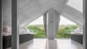 Een moderne zolderkamer met een schuine kap, twee bedden en een groot raam met uitzicht op een bebost landschap.