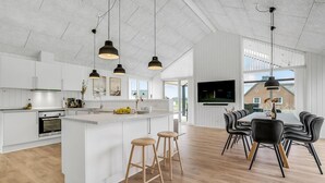 Een moderne keuken met witte kasten, een centraal eiland met krukken, een eethoek met zwarte stoelen, en een aan de muur bevestigde televisie.