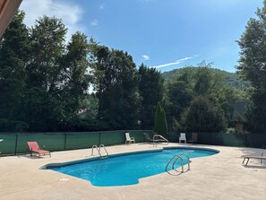 Une piscine avec une plage en béton, entourée d’arbres et d’une clôture verte.
