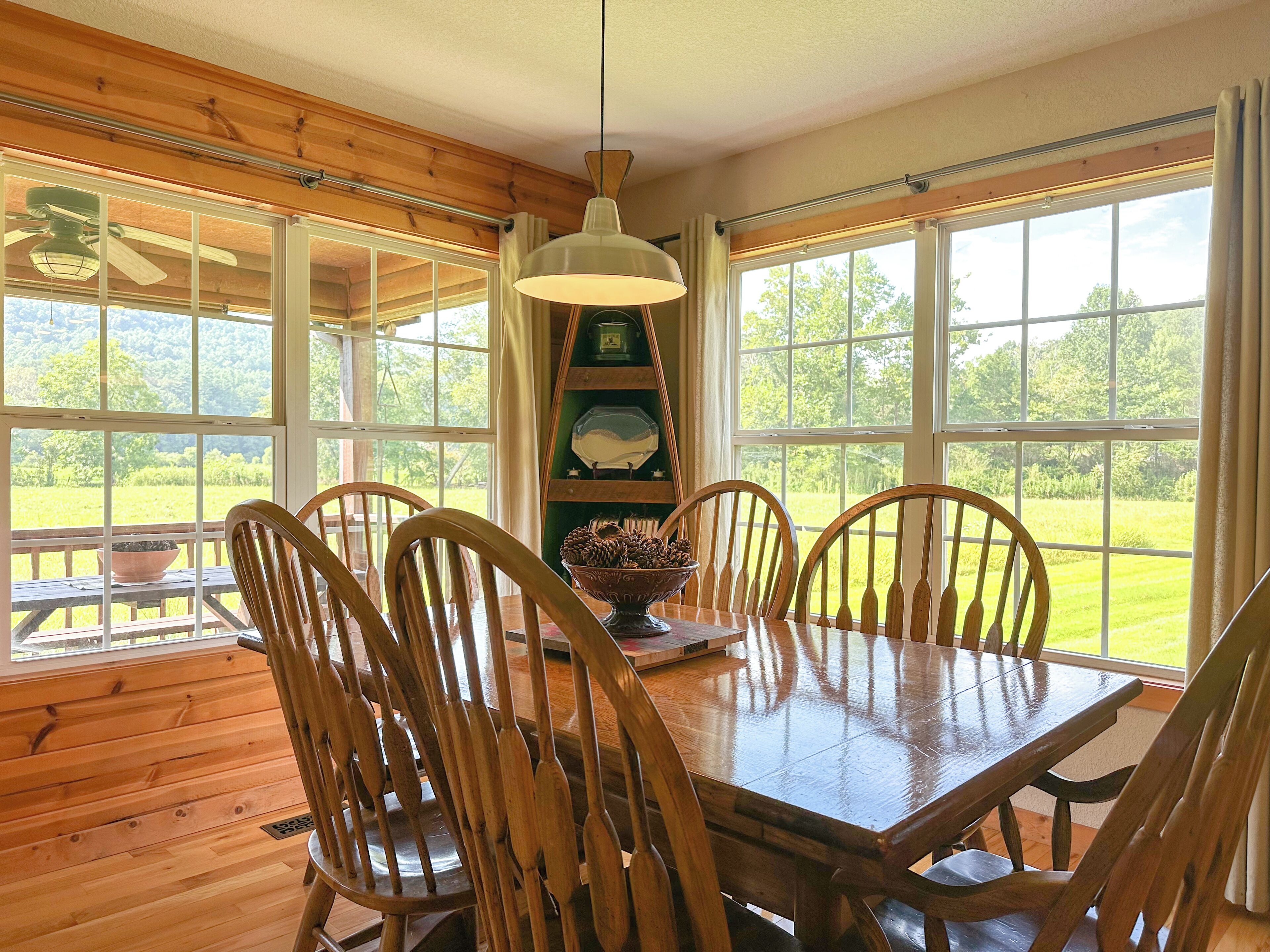 A wooden dining table with six chairs, a hanging light fixture, and a view of a green landscape through large windows.