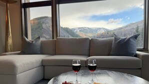 Un salon moderne doté d’une grande fenêtre, d’un canapé gris et d’une table basse ronde sur laquelle se trouvent un verre de vin et des amuse-gueules.