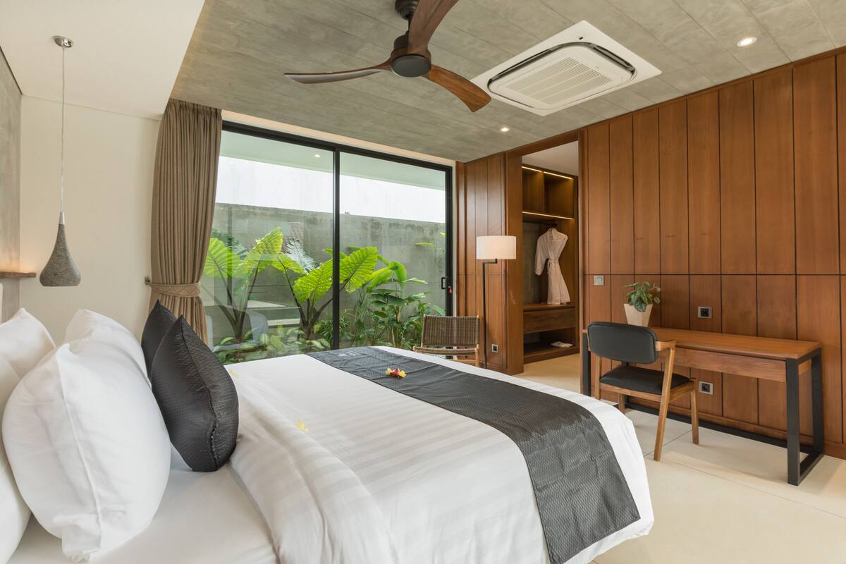 A modern hotel room with a large bed, a desk, and a view of greenery outside.