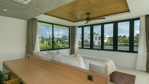 A modern hotel room with a large bed, a wooden desk, and a view of greenery and buildings through large windows. - Villa NVL Canggu by Nagisa Bali (Canggu)