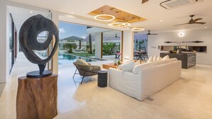 A modern living room with a white sofa, a wicker chair, and a coffee table. There is a large abstract sculpture on a wooden base. The room has a ceiling fan, recessed lighting, and a view of a swimming pool and outdoor area. - Villa NVL Canggu by Nagisa Bali (Canggu)