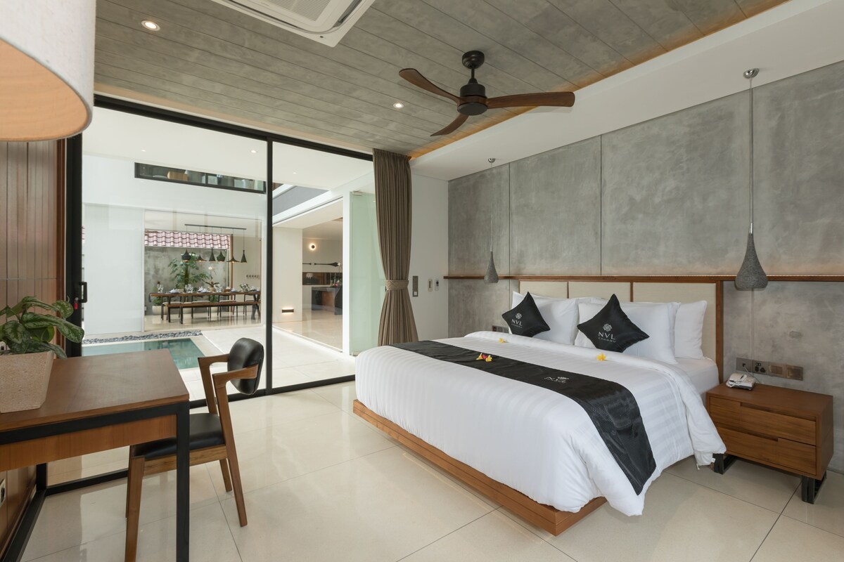 A modern hotel room with a large bed, a dining area with a table and chairs, a ceiling fan, and a view of a restaurant and pool area.