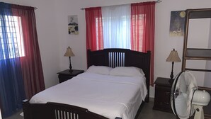 Een net opgemaakt bed met witte lakens, een houten hoofdbord, twee nachtkastjes met lampen, een staande ventilator en gordijnen in rood en wit.