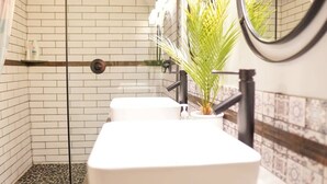 Un baño con paredes de azulejos blancos, una zona de ducha, un lavamanos blanco y una planta en maceta.