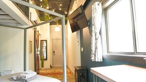 A modern hotel room with a bed, a flat-screen TV mounted on the wall, a window with curtains, and a small table.