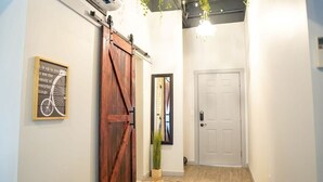 Un pasillo con una puerta de madera rústica, una cita enmarcada en la pared, una planta en maceta y una puerta blanca al final.