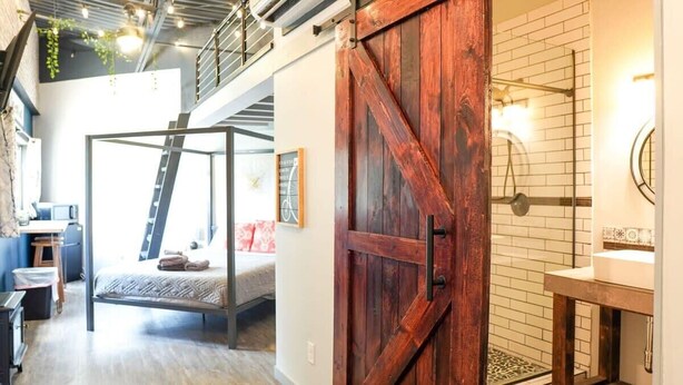 Un dormitorio estilo loft con una escalera metálica, un gran puerta de madera y un baño con lavamanos blanco y salpicadero de azulejos.