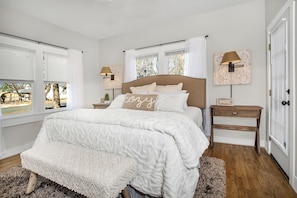 Une chambre à coucher avec un grand lit, deux tables de chevet, une commode, une fenêtre avec des stores et une porte dotée d’une fenêtre.