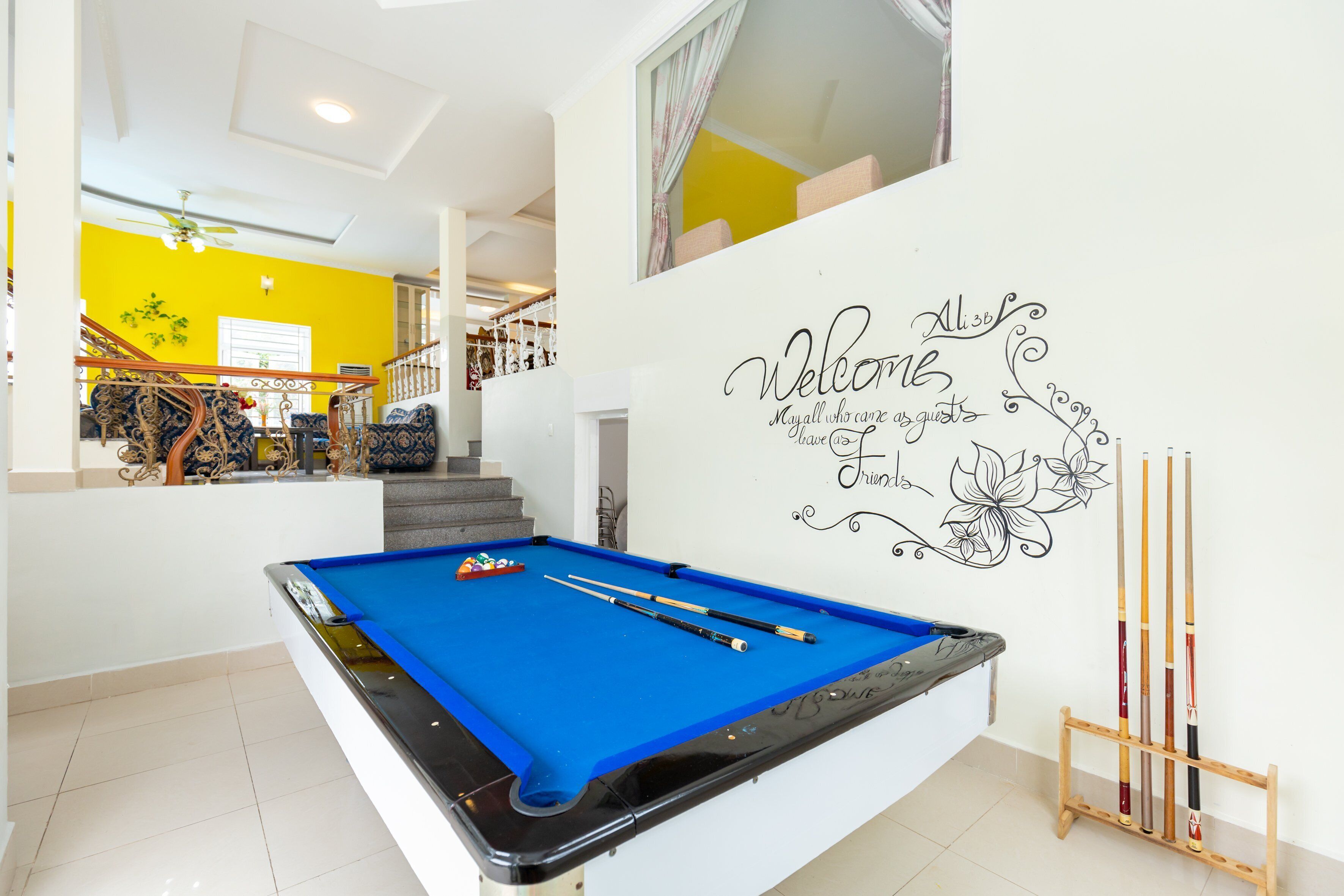 A pool table with pool cues, a staircase, and a welcome sign on the wall.