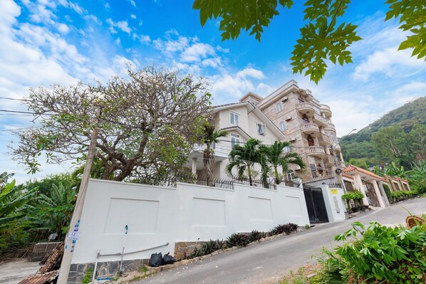 A large, multi-story building with balconies, surrounded by lush greenery and a clear sky.