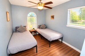 Une chambre avec deux lits simples, un ventilateur de plafond, une fenêtre avec des stores et une petite table avec une lampe.