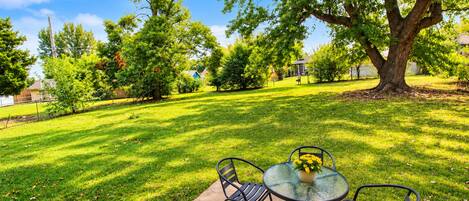 A backyard with a glass table and chairs, surrounded by a well-maintained lawn and trees.