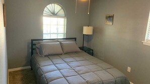 Un dormitorio con cama, ventilador de techo, ventana con persianas, mesita de noche con lámpara y un cuadro en la pared.