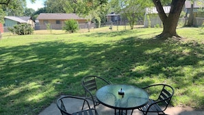 Un patio con una mesa de vidrio y cuatro sillas, rodeado por un césped y árboles.