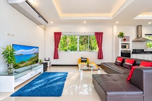 A modern living room with a sofa, a coffee table, a TV, and a kitchen area. - 4 Br Private Pool Villa GA (Chalong)