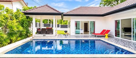 Una piscina con solárium enlosado, un porche cubierto con mobiliario y un ventanal de vidrio que da a una habitación.