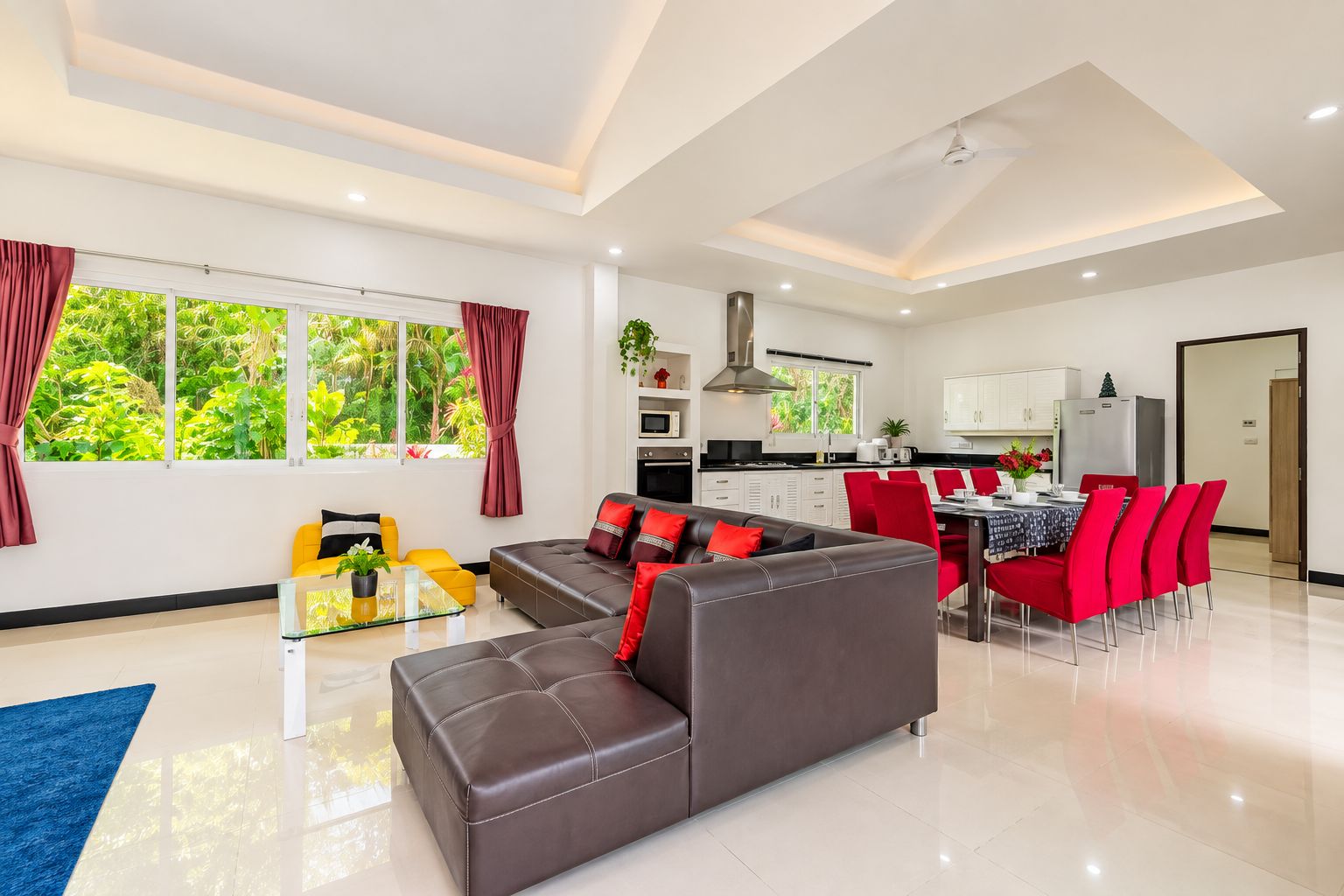 Un salon moderne avec un canapé, un coin repas avec des chaises rouges, une cuisine avec des meubles blancs et de grandes fenêtres avec des rideaux.