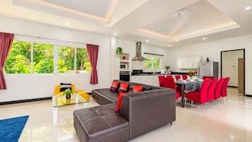 Un salon moderne avec un canapé, un coin repas avec des chaises rouges, une cuisine avec des meubles blancs et de grandes fenêtres avec des rideaux.