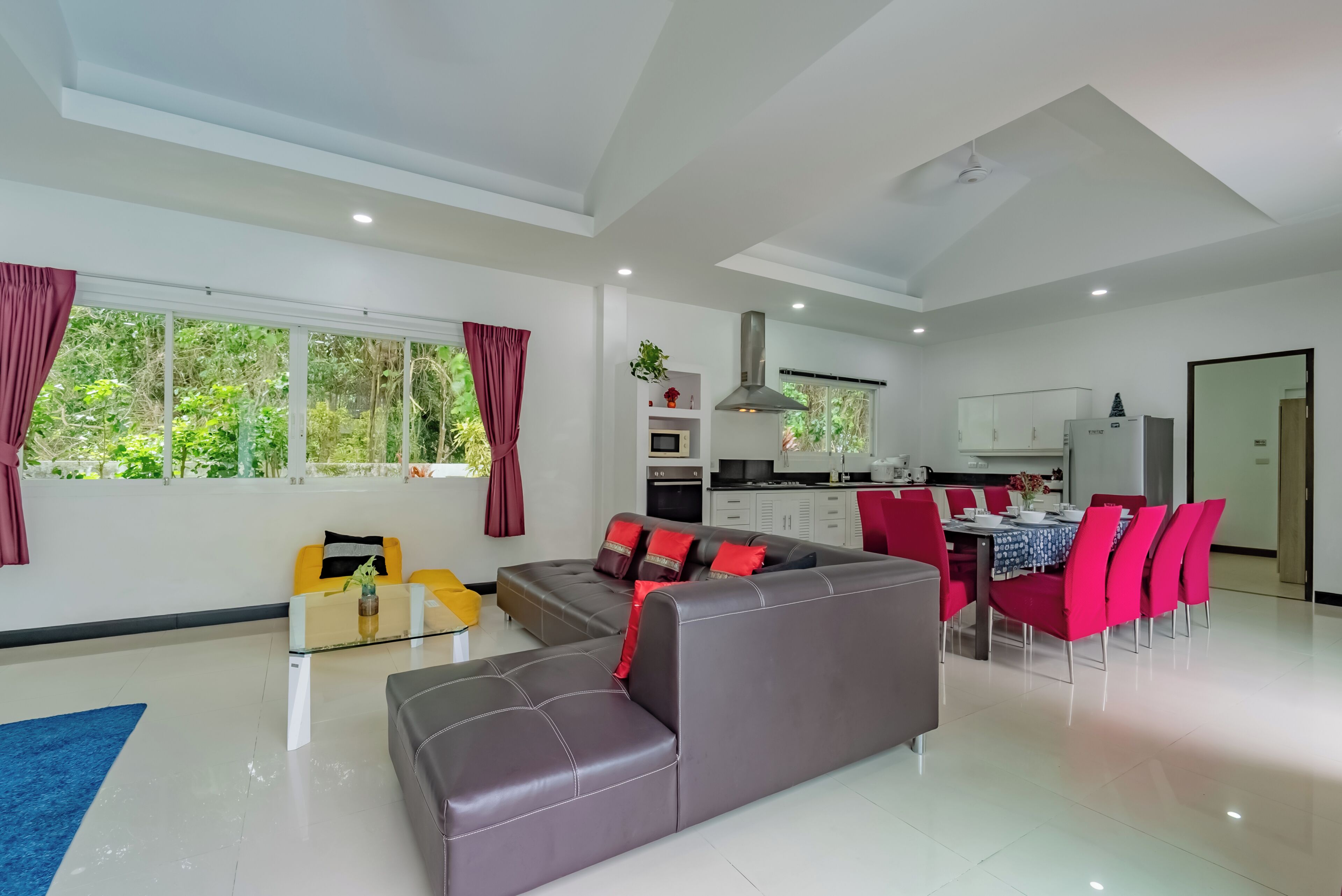 A modern living room with a sofa, dining table, and chairs, a kitchen in the background, and large windows with curtains.