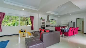 A modern living room with a sofa, dining table, and chairs, a kitchen in the background, and large windows with curtains.