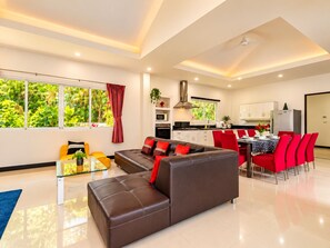 A modern living room with a sofa, a glass coffee table, and a dining area with red chairs. - 4 Br Private Pool Villa GA (Chalong)