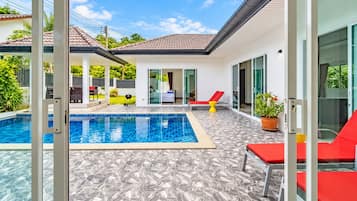 Un área de piscina exterior moderna con solárium empinado, sillas de descanso rojas y una puerta de cristal que da a un porche cubierto.