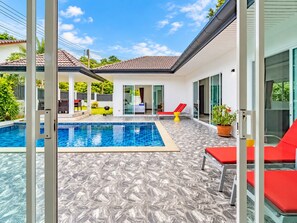 Un área de piscina exterior moderna con solárium empinado, sillas de descanso rojas y una puerta de cristal que da a un porche cubierto.